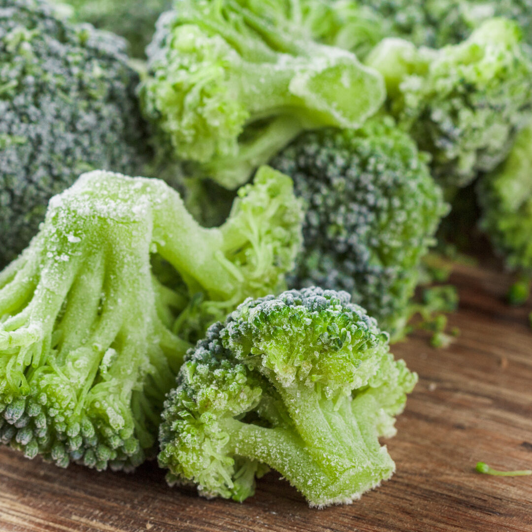 Speciality Frozen Vegetables – Our Offering – Frozen Brands Close-up view of frozen broccoli florets scattered onto a wooden table.