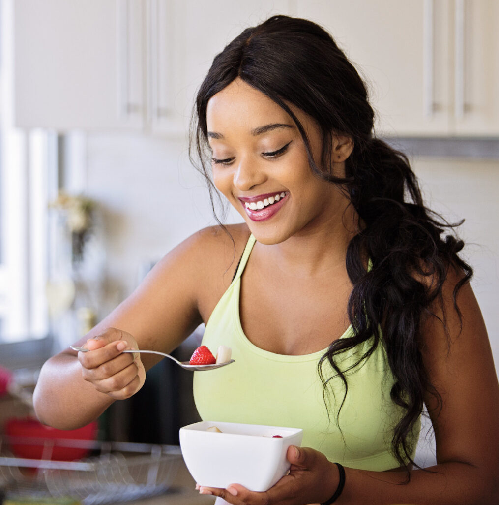 Smiling woman holding a bowl of fruit in her one hand, while using her other lifting a spoon to her mouth.